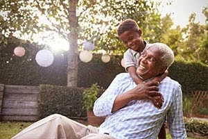 Smiling grandfather playing with grandson in a sunlit backyard with hanging lanterns.