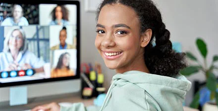 Smiling young woman in a hoodie on a video call with multiple participants on her computer screen.