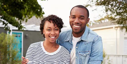 Happy couple outdoors, smiling in front of a house; the woman wears a striped shirt, and the man a denim jacket.