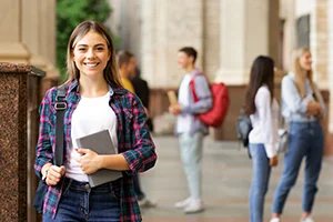 Smiling female student holding a tablet and backpack, standing outside a school building with other students in the background.