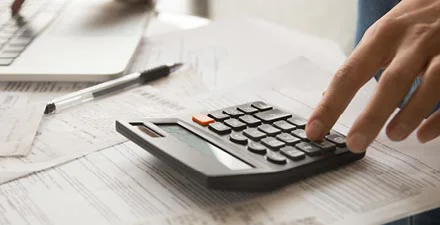 Close-up of hand using a calculator on top of financial documents next to a laptop.