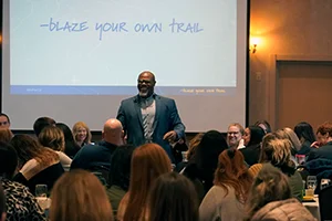 Speaker presenting to an audience with "Blaze Your Own Trail" on the screen behind them.