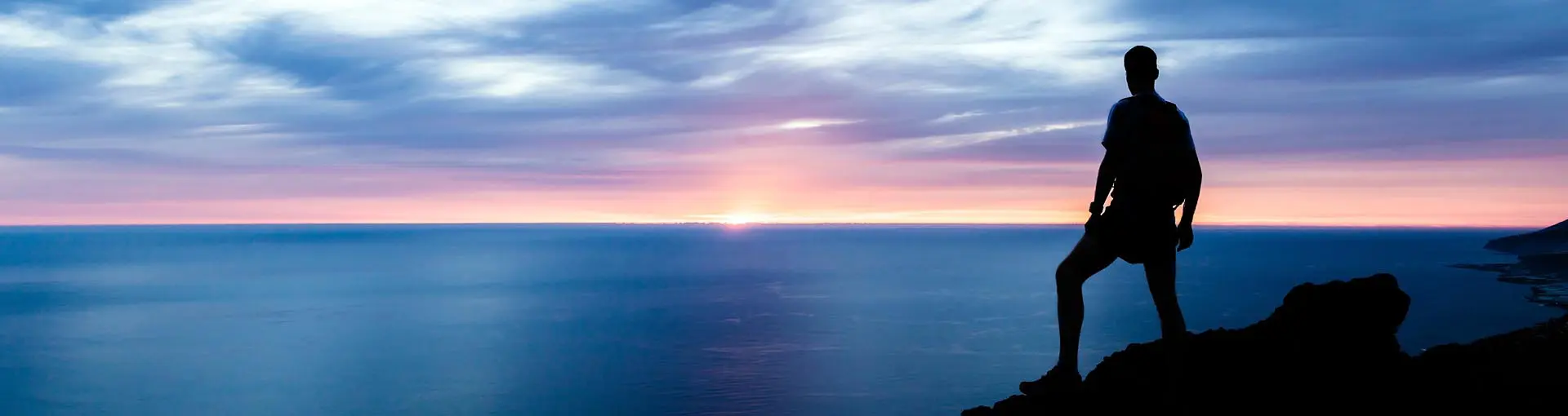 Silhouette of person standing on cliff overlooking ocean at sunset.
