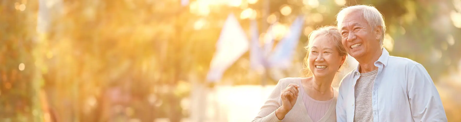 Smiling older couple walking outdoors in warm sunlight with trees in the background.
