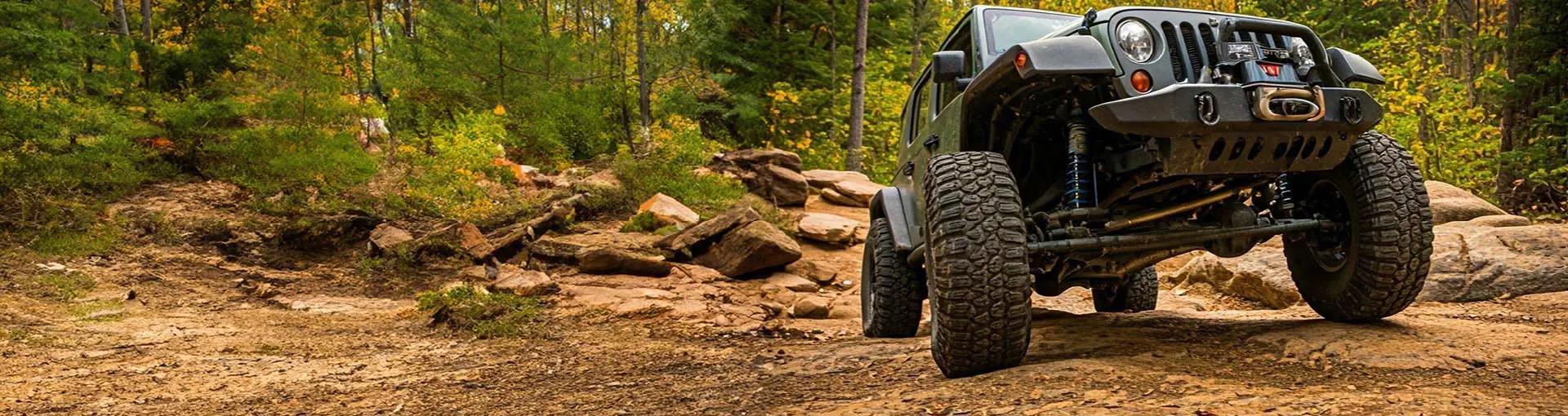 Jeep driving off-road through a rocky forest trail surrounded by trees and bushes.