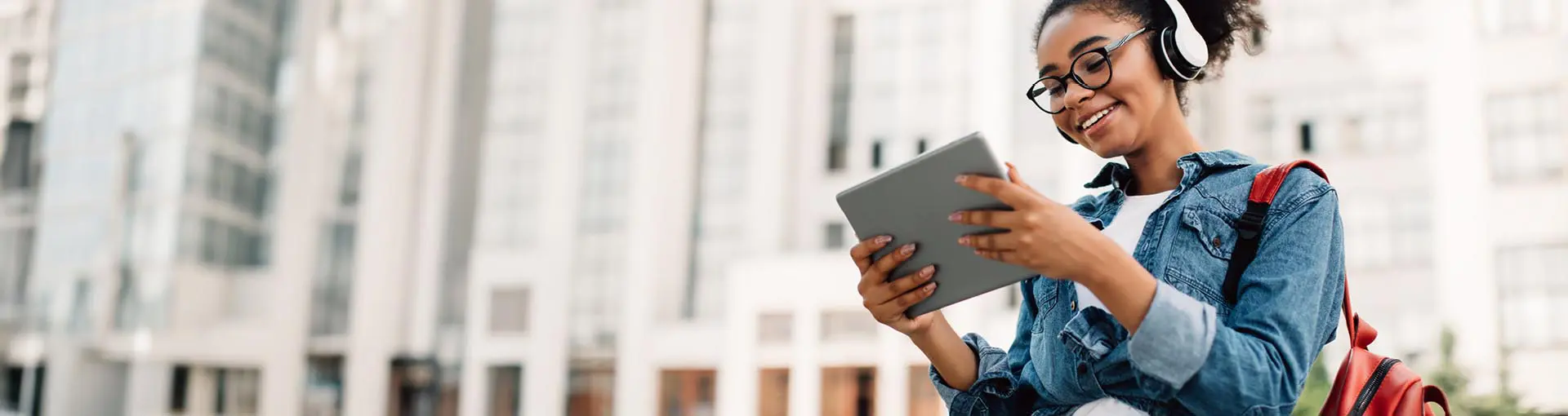 Smiling young woman with headphones using a tablet outdoors in front of tall buildings.