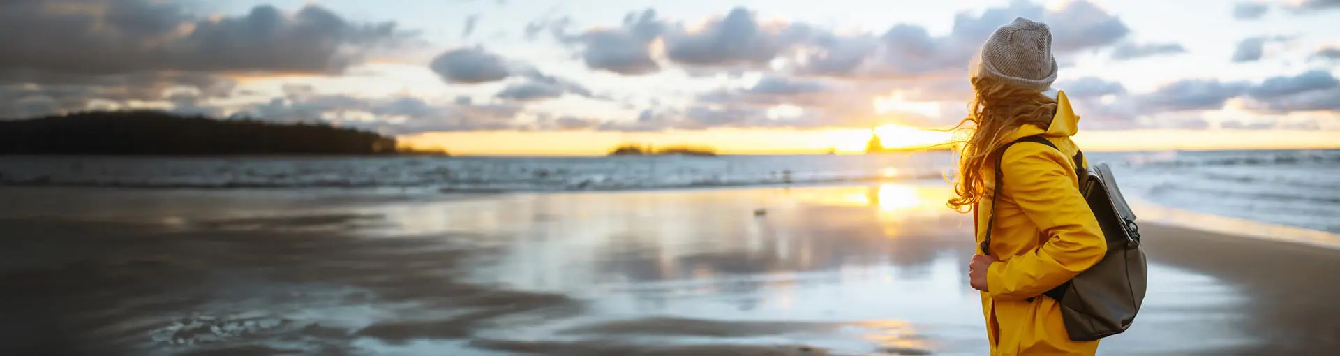 Woman in a yellow rain jacket with a backpack looking at the sunset on the beach.
