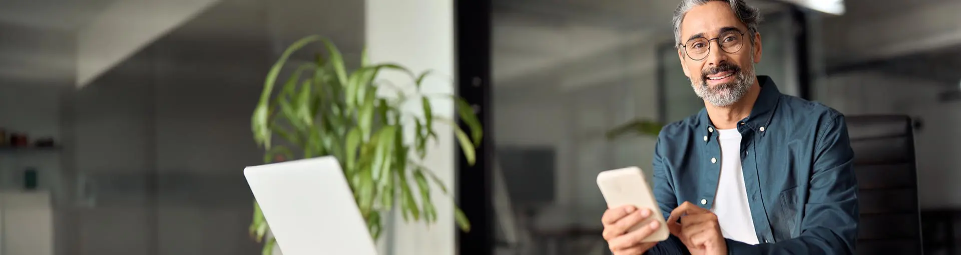 Smiling man with glasses using a smartphone at a desk with a laptop and plant in the background.