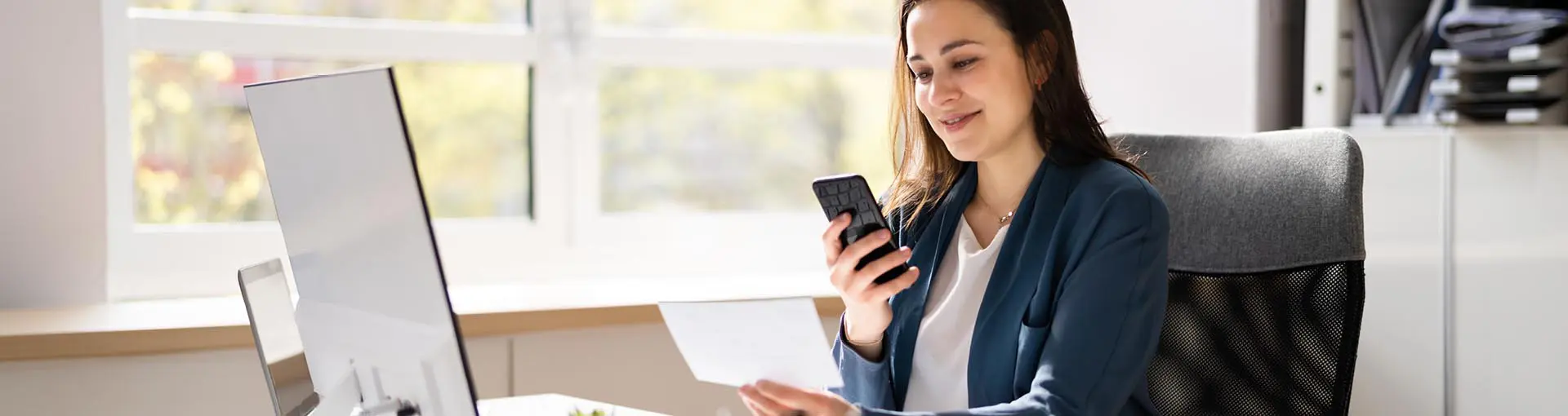 Woman sitting at a desk holding a phone and looking at a document with a laptop in front of her.