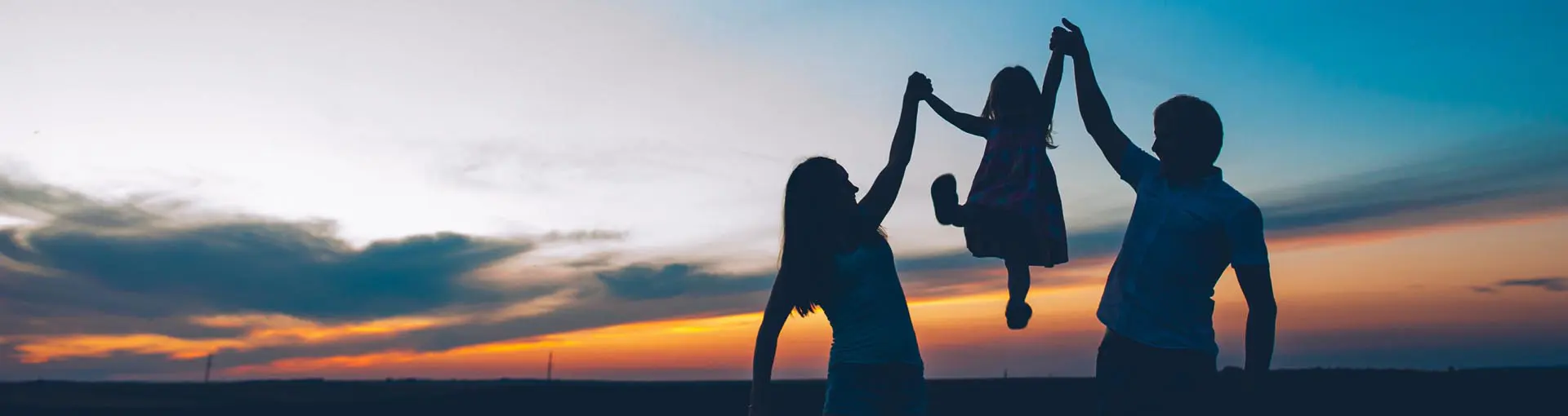 Silhouette of parents lifting their child in the air at sunset with a scenic sky in the background.